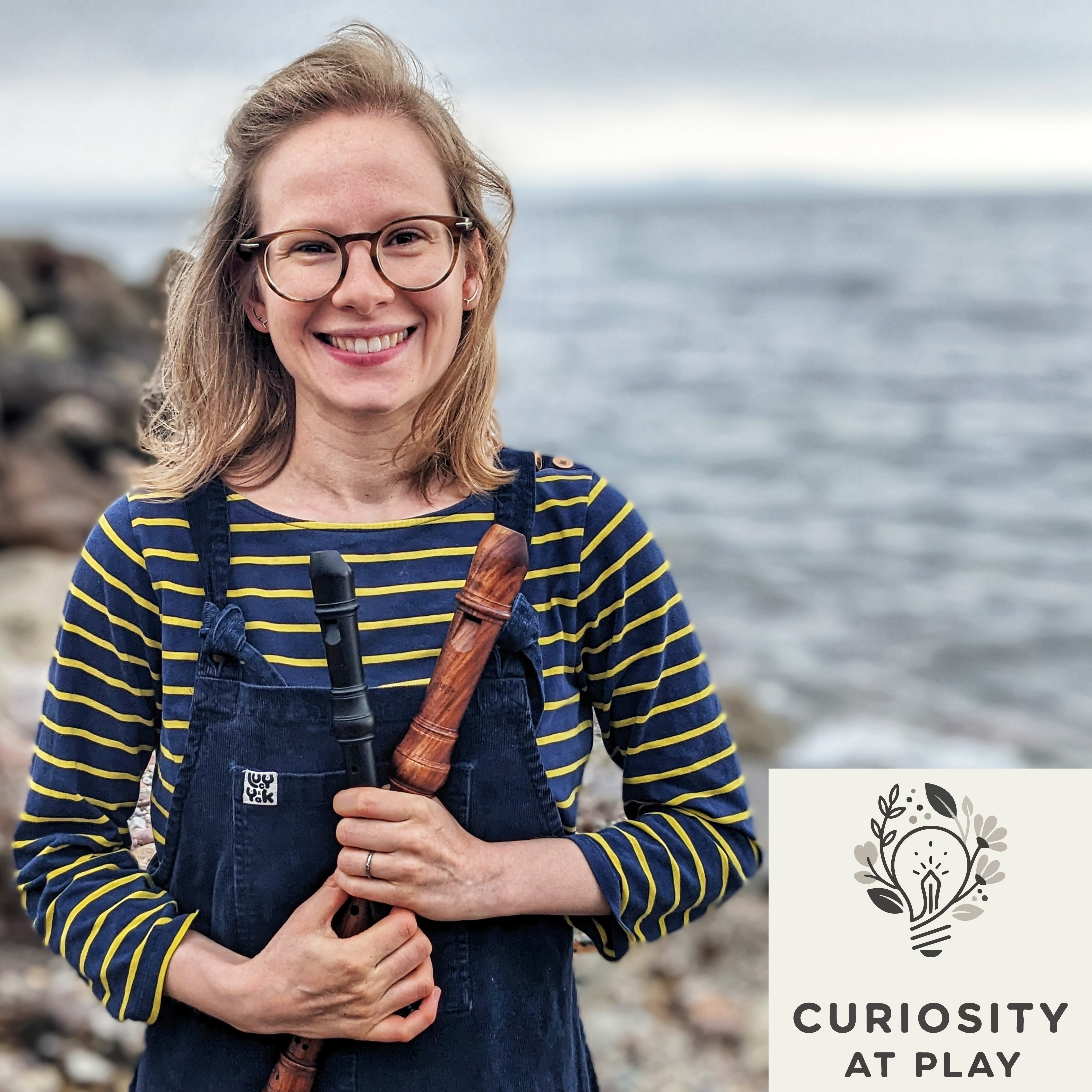 Smiling woman in overalls holding two recorders by the ocean with Curiosity at Play logo.