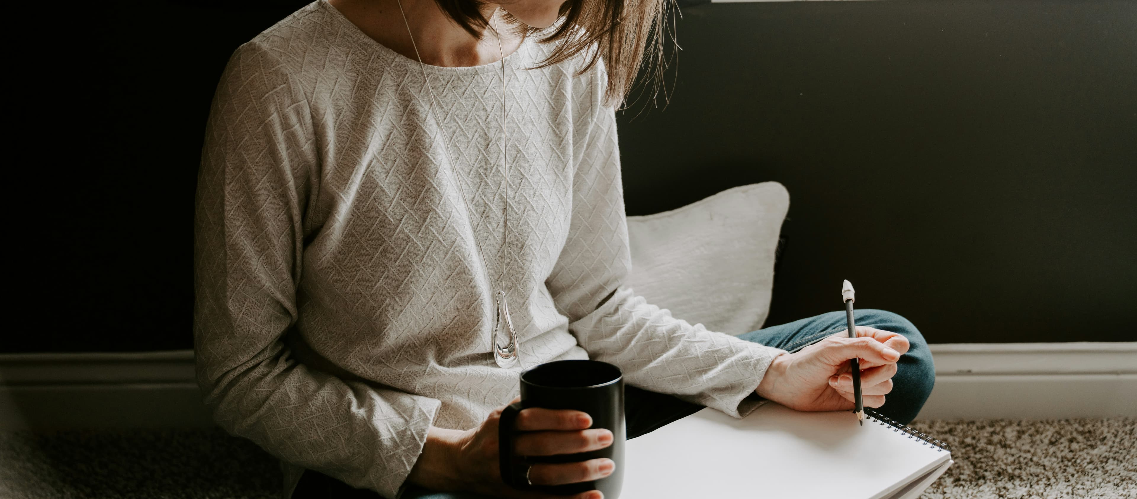 Person in a textured sweater sits on the floor, holding a black mug and writing.
