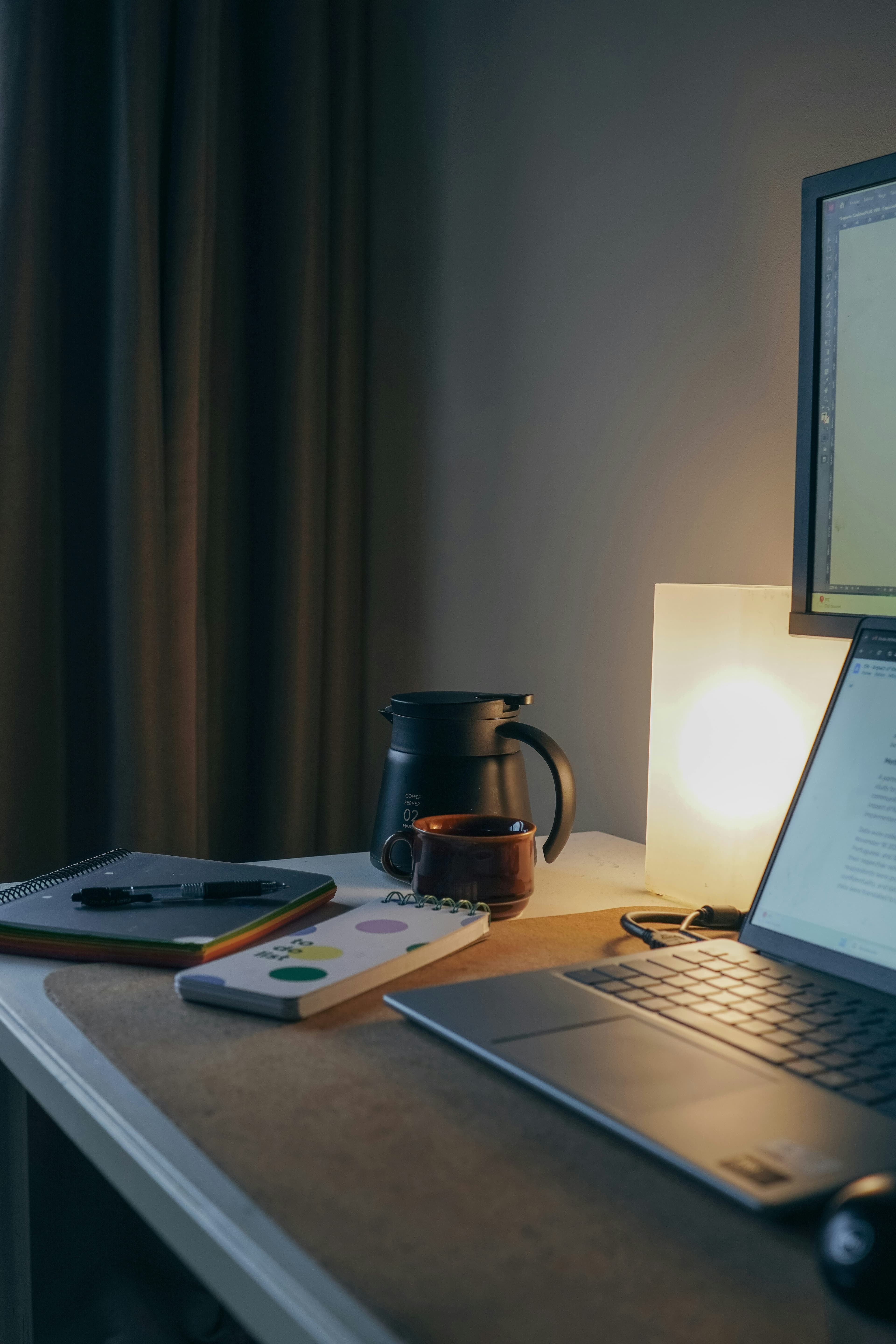Laptop, coffee carafe, and notebooks on a desk illuminated by a warm glowing lamp.