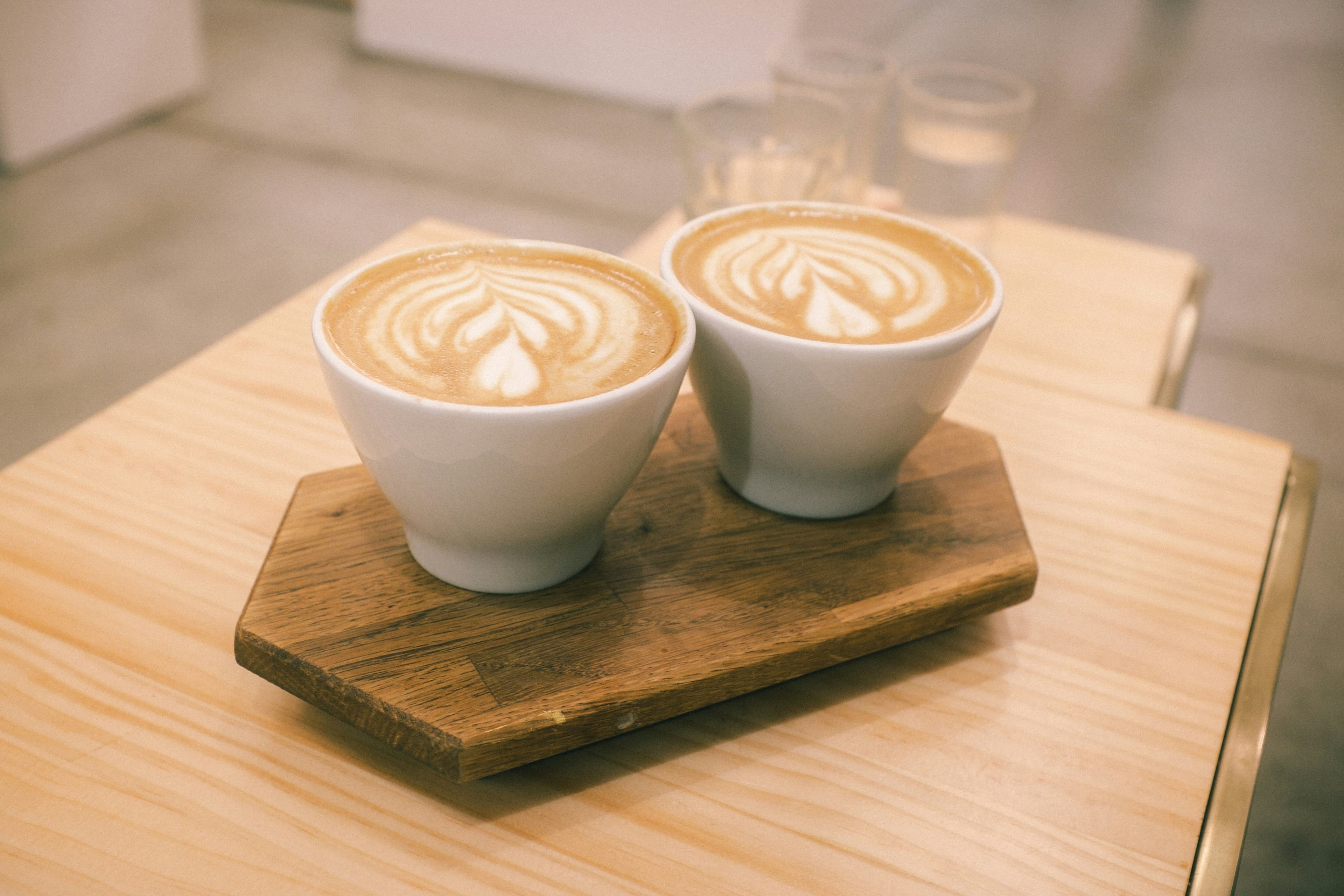Two white cups of latte with leaf latte art on a wooden serving board.