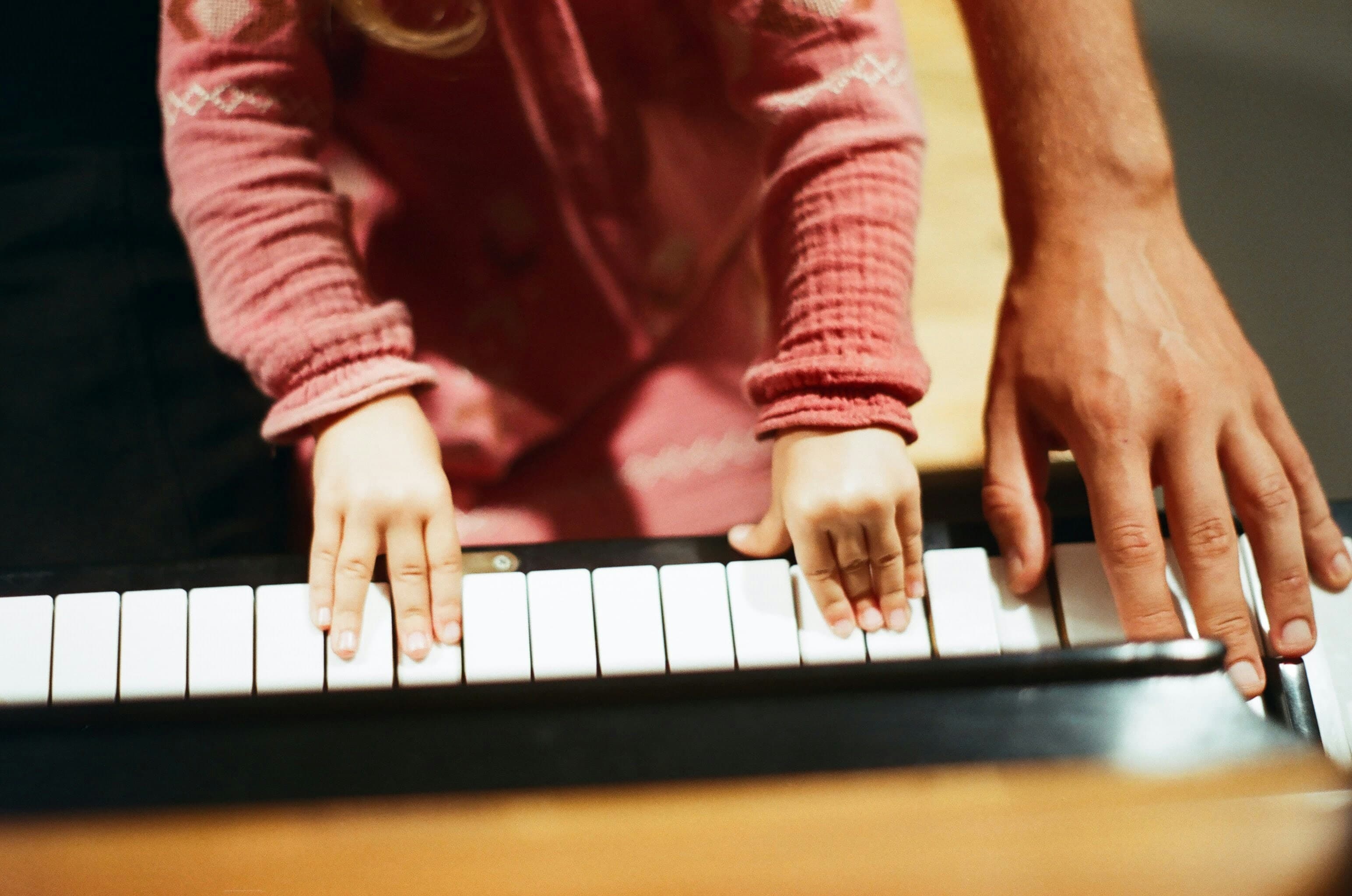 A child in a pink sweater plays a toy piano beside an adult's hand.